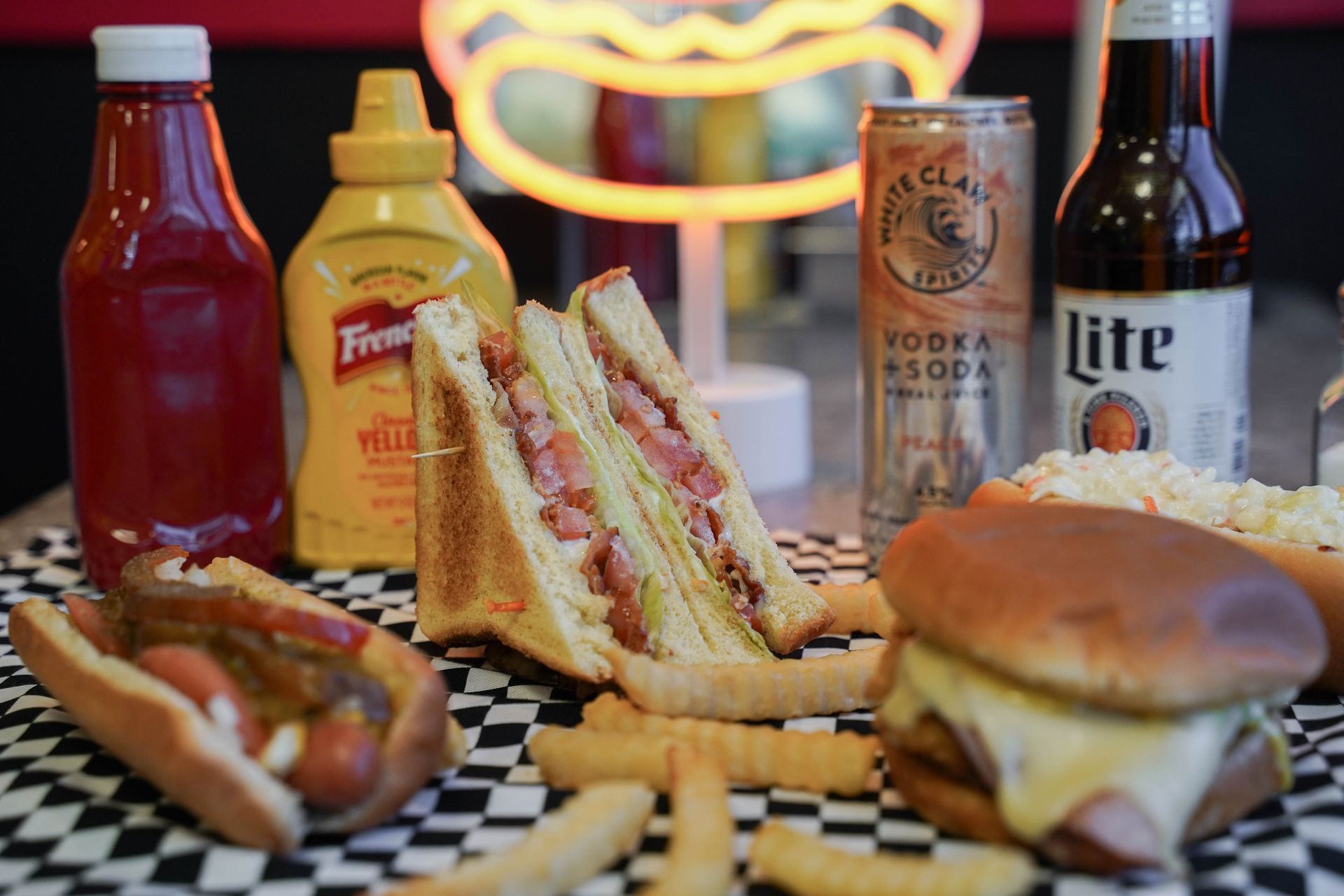 Assorted fast food and condiments on a checkered tablecloth with beer bottles in the background
