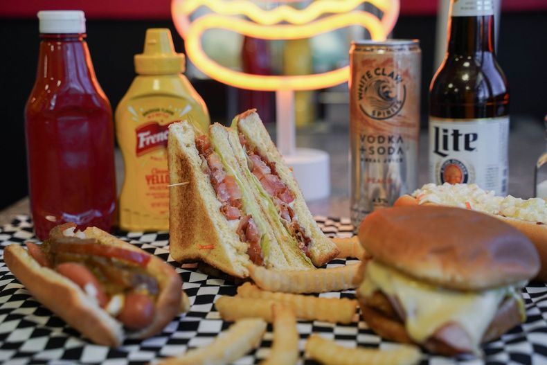 Assorted fast food and condiments on a checkered tablecloth with beer bottles in the background