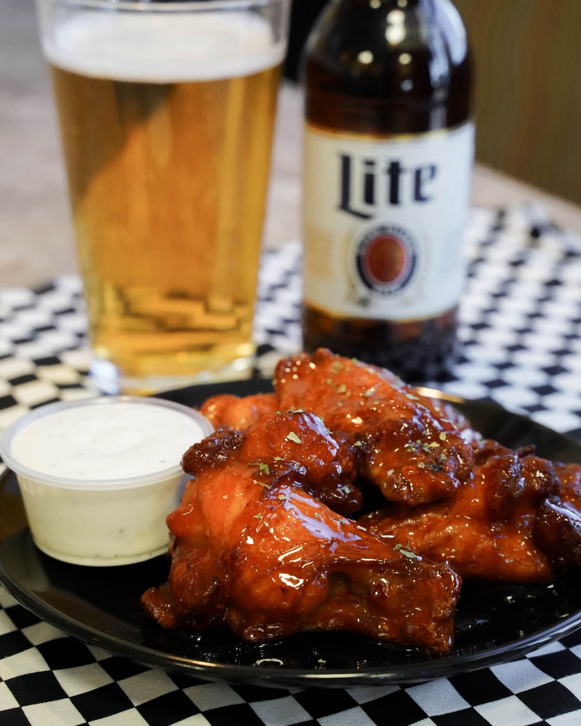 Plate of saucy wings with dipping sauce, a beer glass, and a Lite beer bottle on a checkered tablecloth