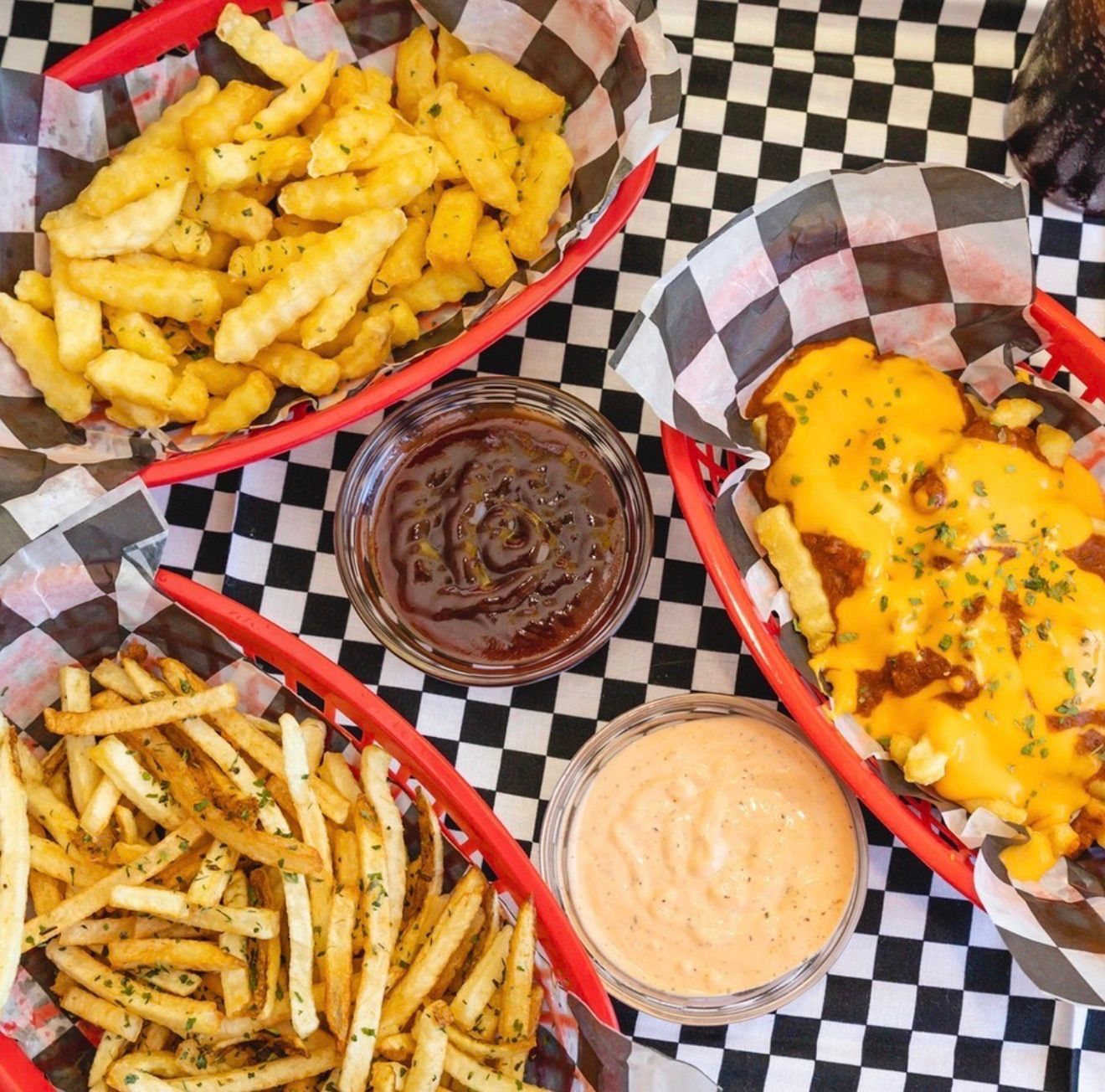 Three baskets of seasoned fries and cheesy fries with two dipping sauces on a checkered tablecloth.