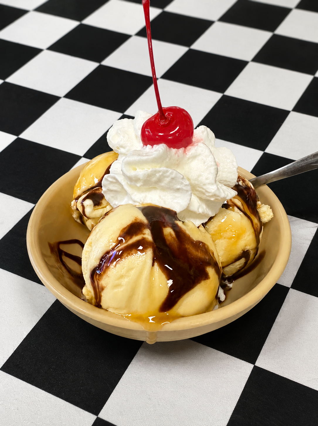 Ice cream sundae with whipped cream, chocolate sauce, and a cherry on a checkered tablecloth