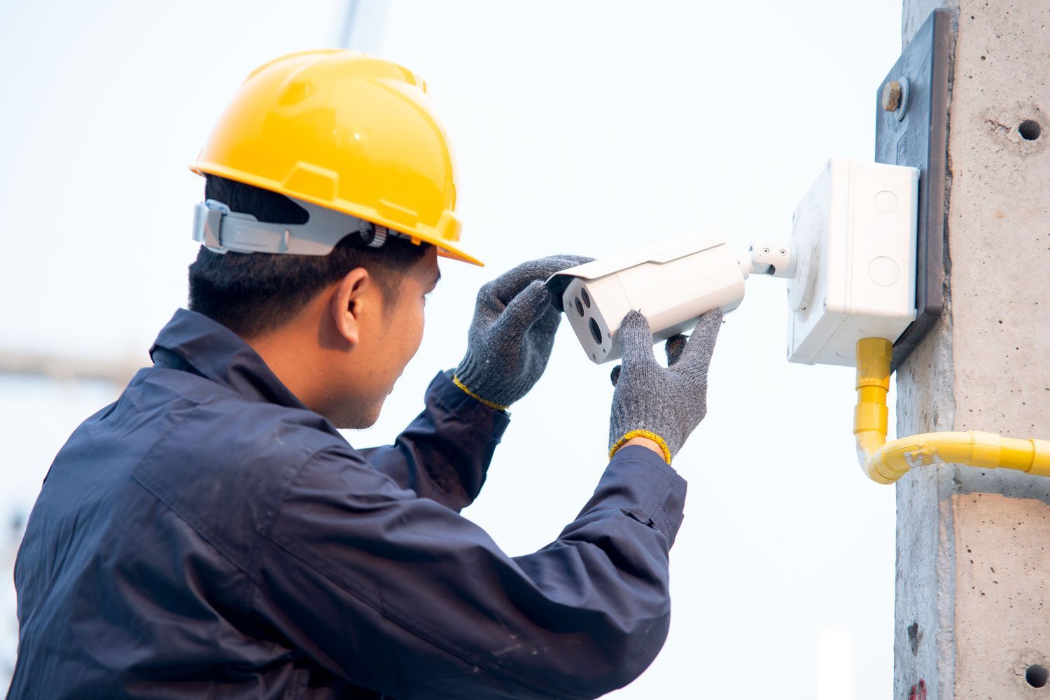Construction worker in a yellow hard hat installing a security camera on a concrete pole.