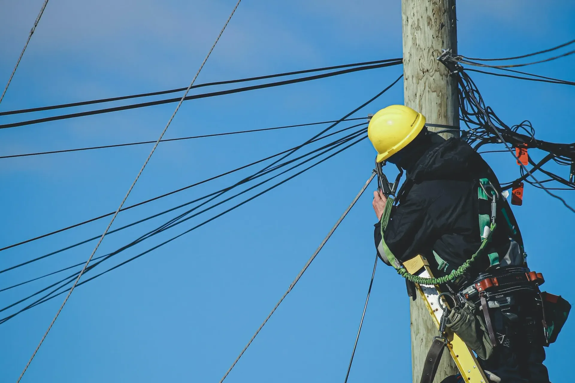 Worker in yellow hard hat climbing a utility pole with overhead power lines against a blue sky