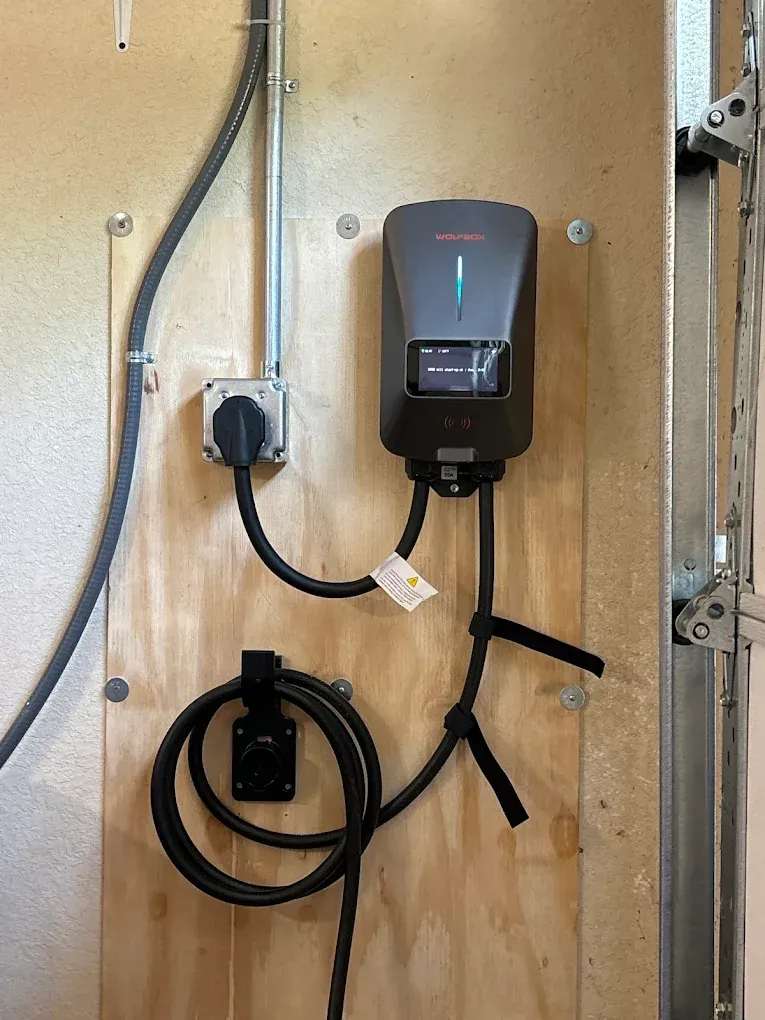 Wall-mounted EV charger with coiled cable and plug on a plywood board in a utility area