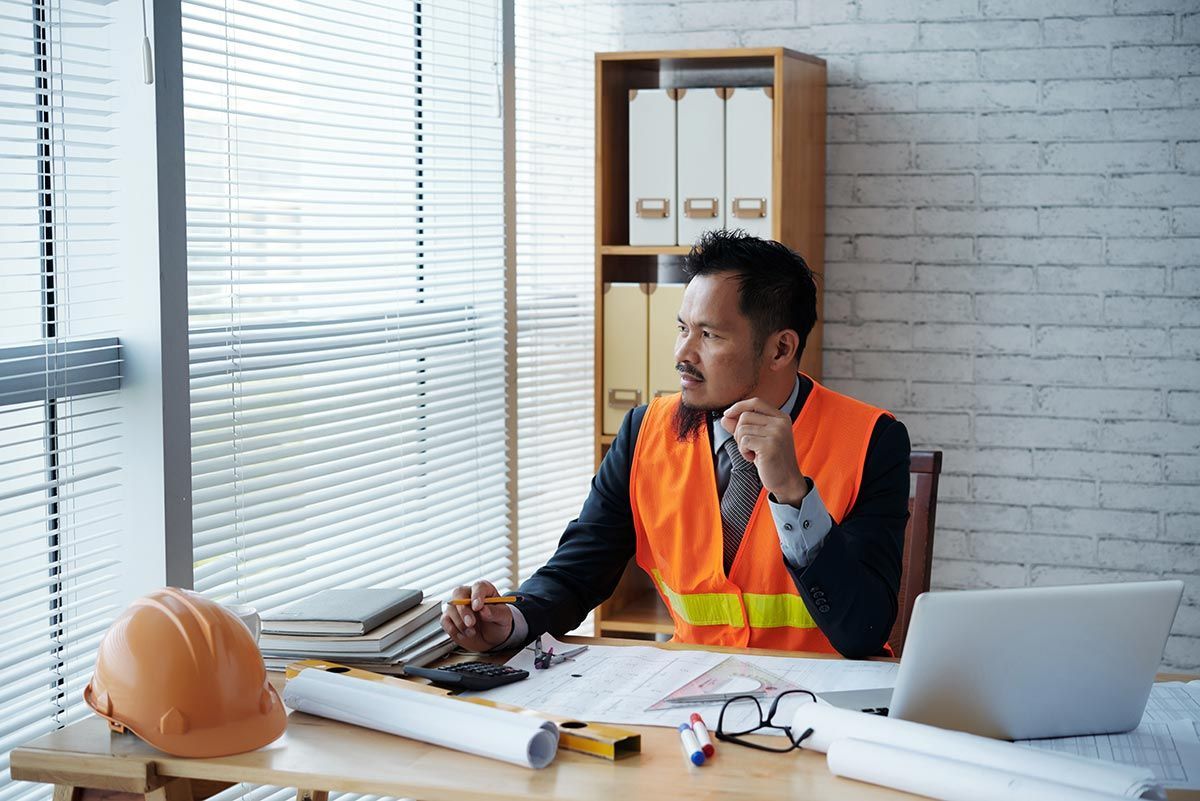Worker in orange safety vest using a phone at a desk with plans, hard hat, and laptop in an office