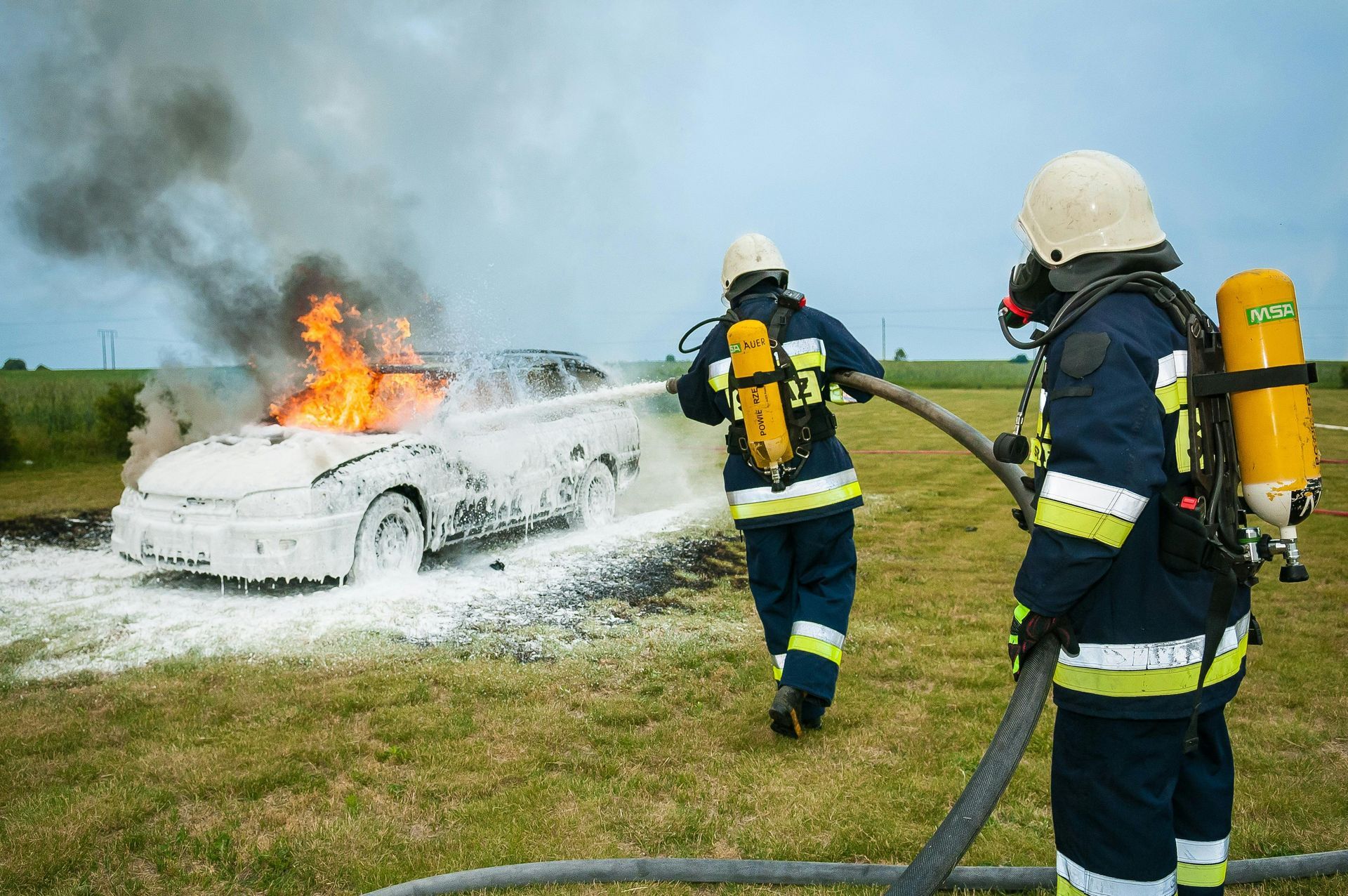 Firefighters extinguishing a car fire with foam in a grassy field.