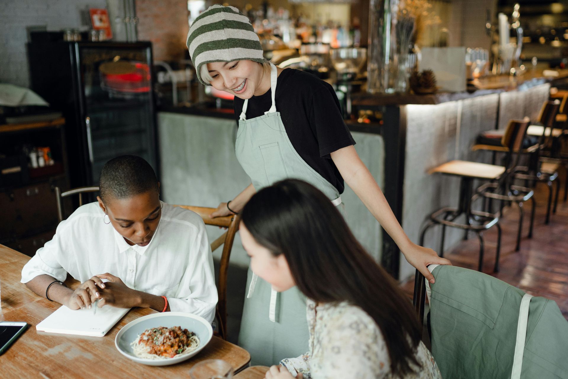 A server in an apron smiling at two customers at a table in a cafe; one is eating.