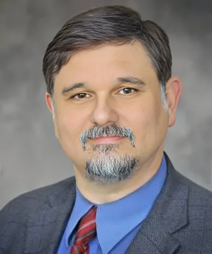 Studio headshot of a man in a gray blazer, blue shirt, and red tie, smiling slightly