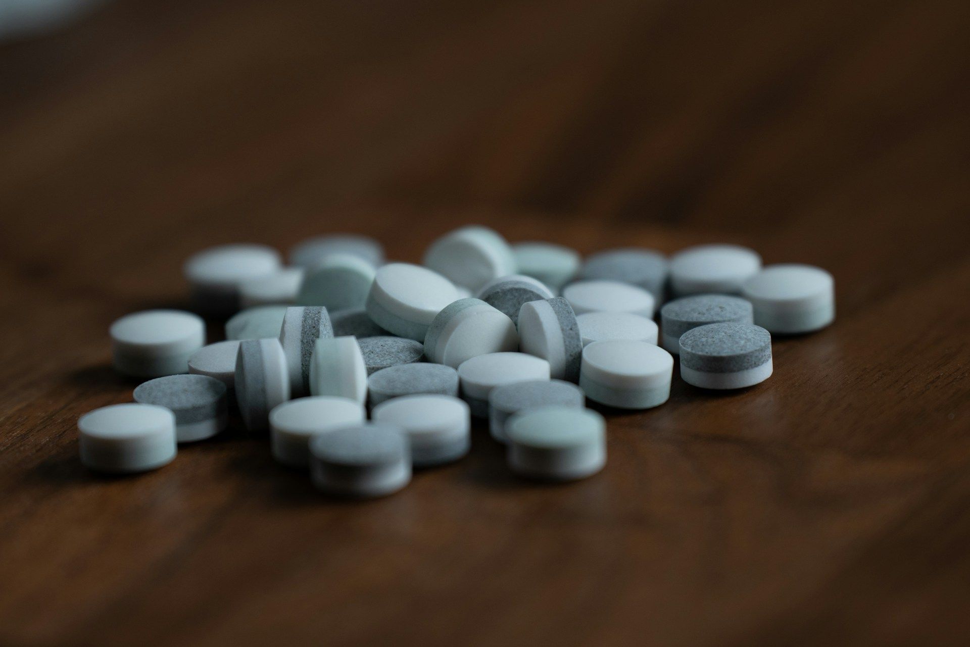 A small pile of round, white and gray pills scattered on a dark wooden surface.
