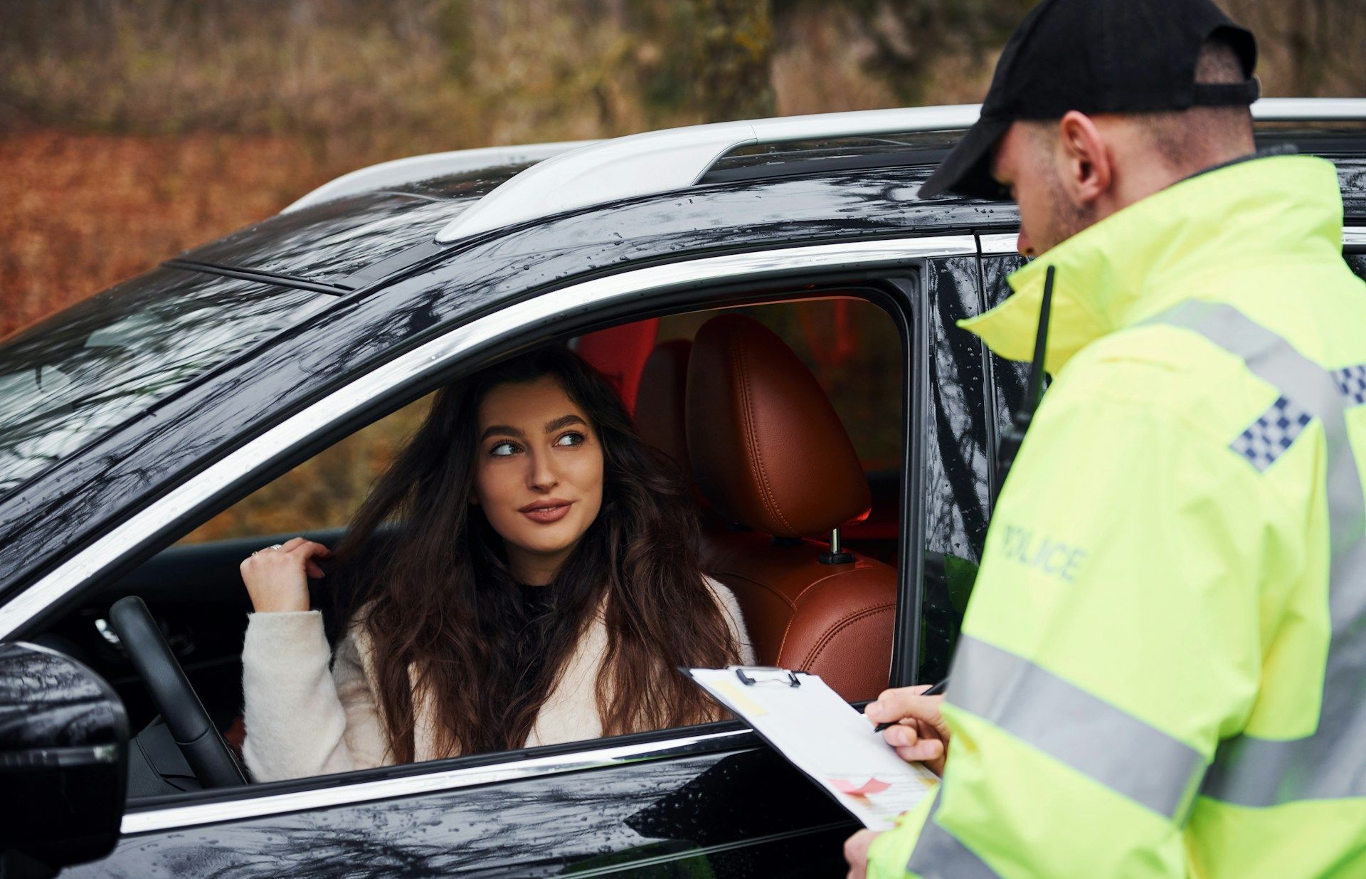 A police officer in a high-visibility jacket stands by a car window, holding a clipboard while talking to the driver.