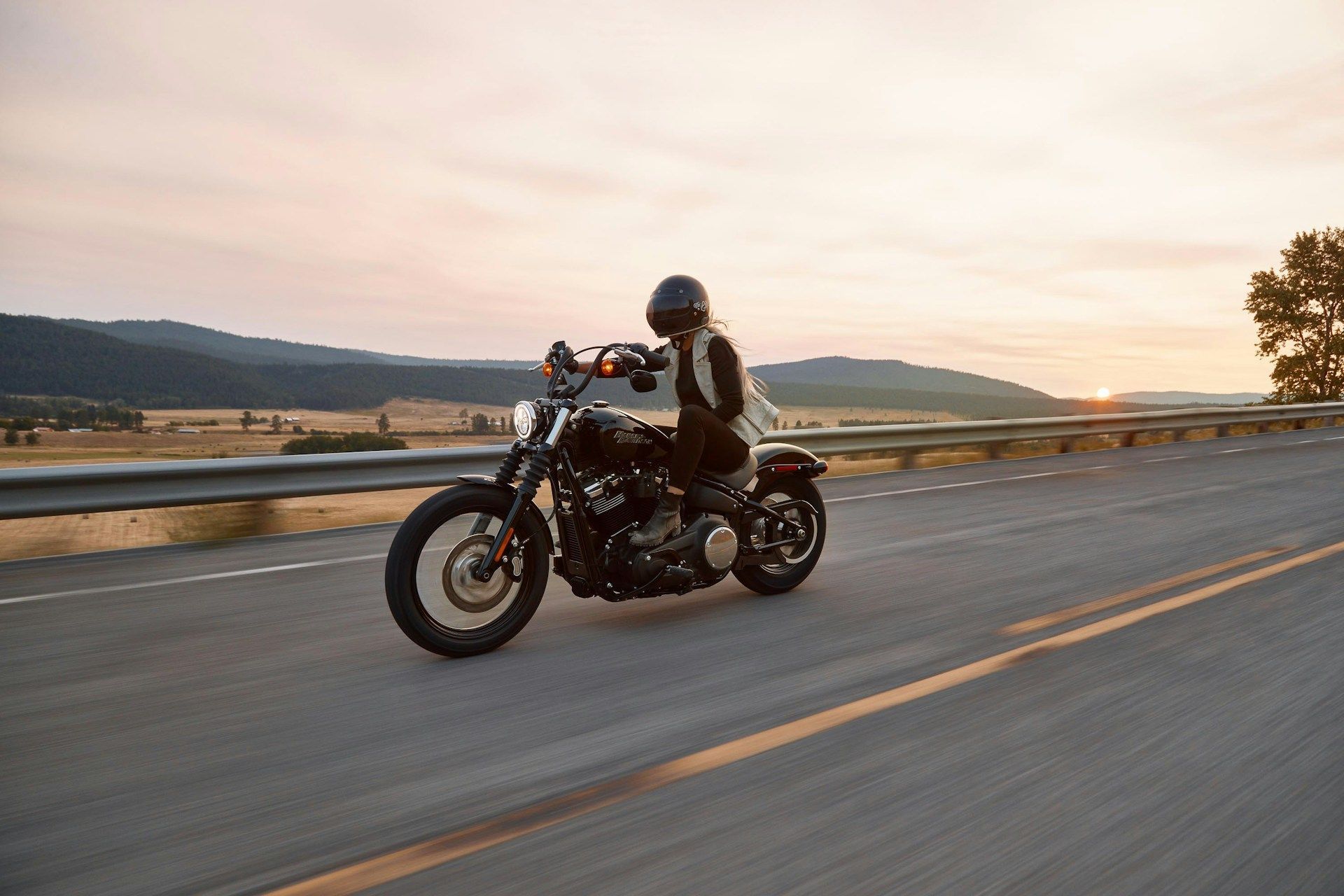A motorcyclist in a black helmet rides a black cruiser motorcycle down an open highway at sunset.