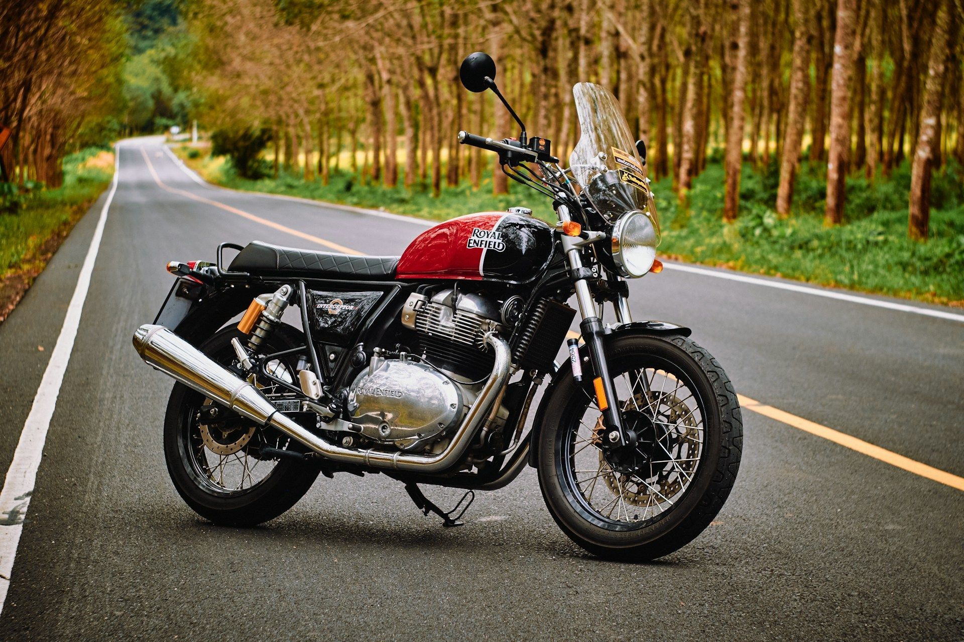 A red and black Royal Enfield motorcycle parked on a paved road lined with trees.