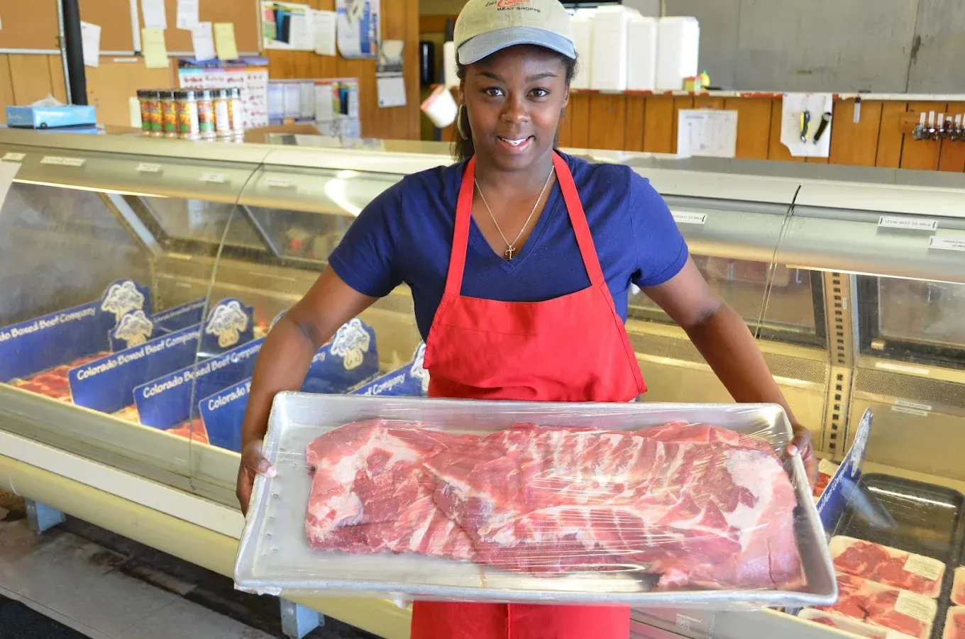 Butcher holding a tray of raw meat in a deli counter, wearing a red apron and cap.