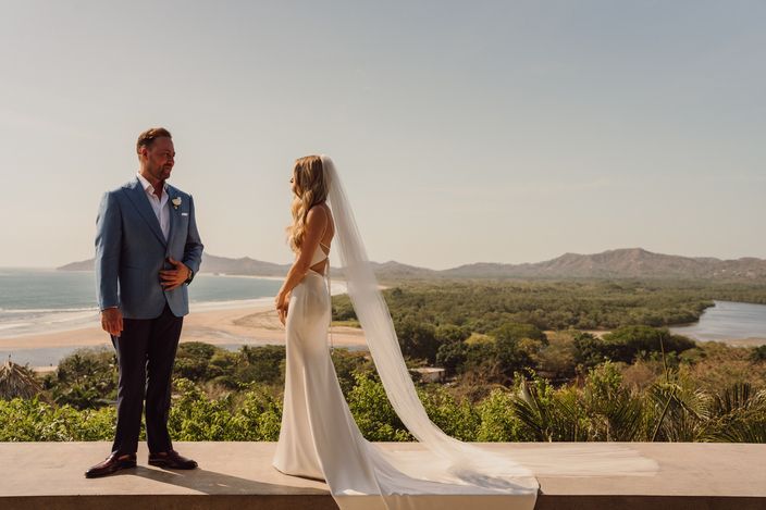 A bride and groom are standing on a balcony overlooking the ocean.