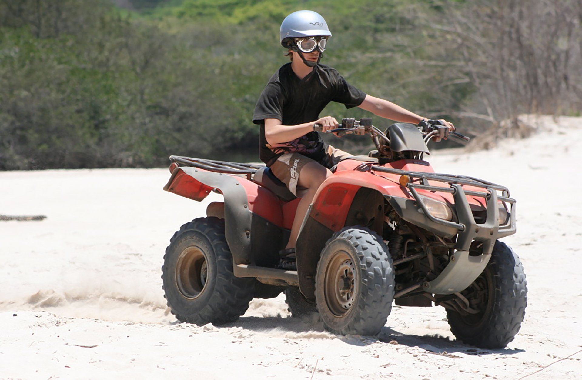 A man wearing a helmet and goggles is riding a red atv on a sandy beach.