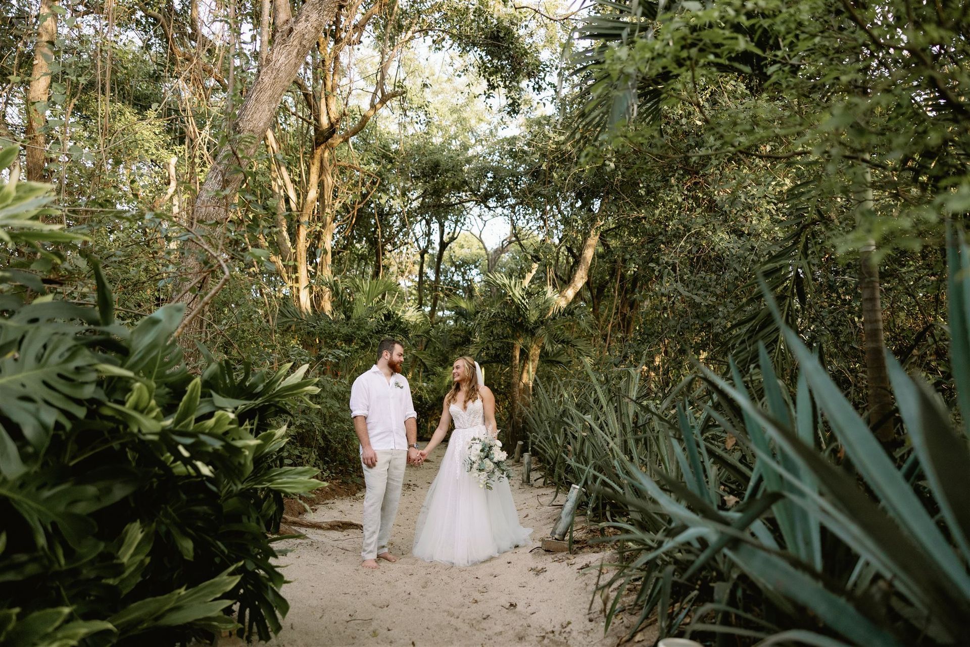 A bride and groom are walking through a forest holding hands.