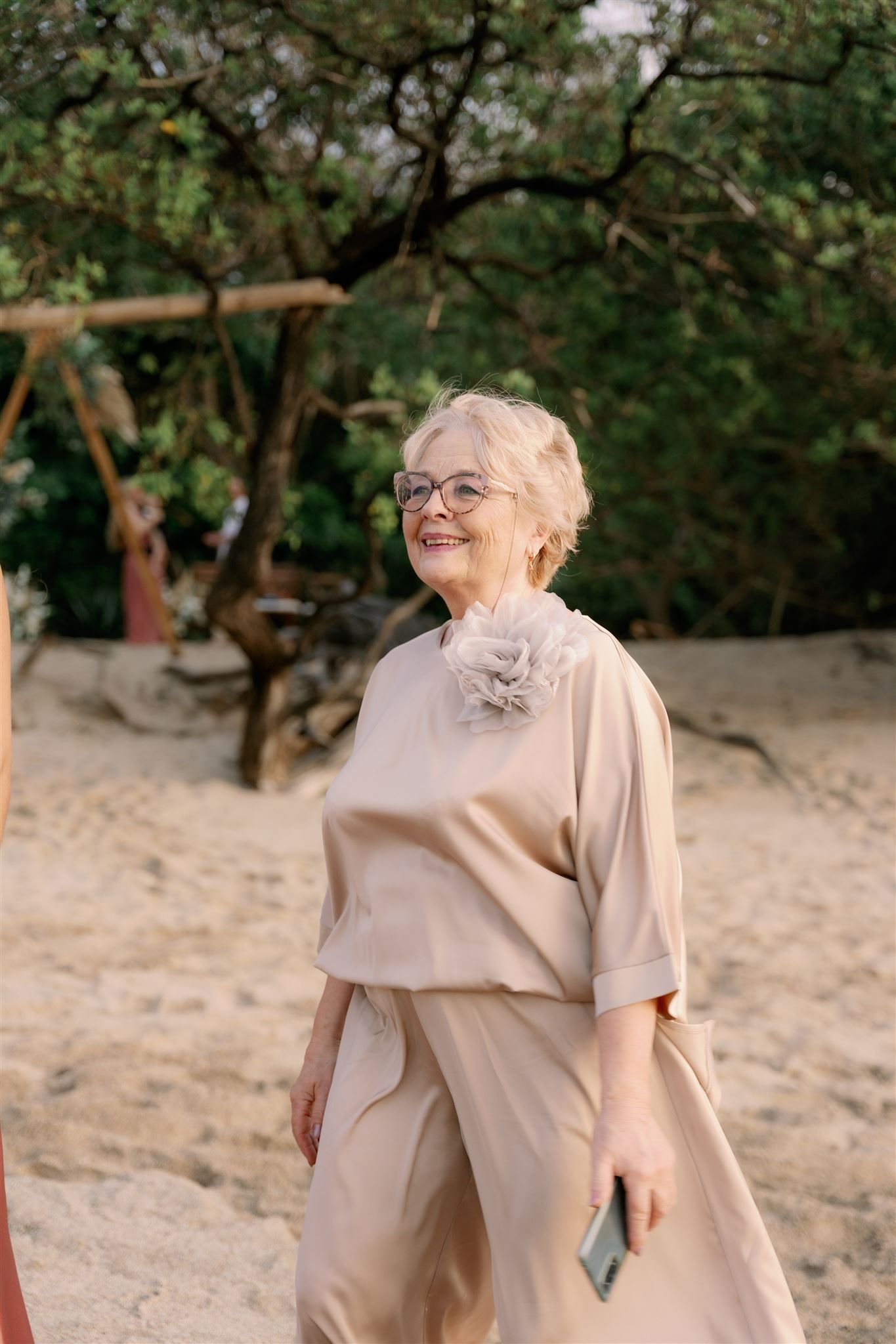 An elderly woman is standing on a sandy beach holding a cell phone.
