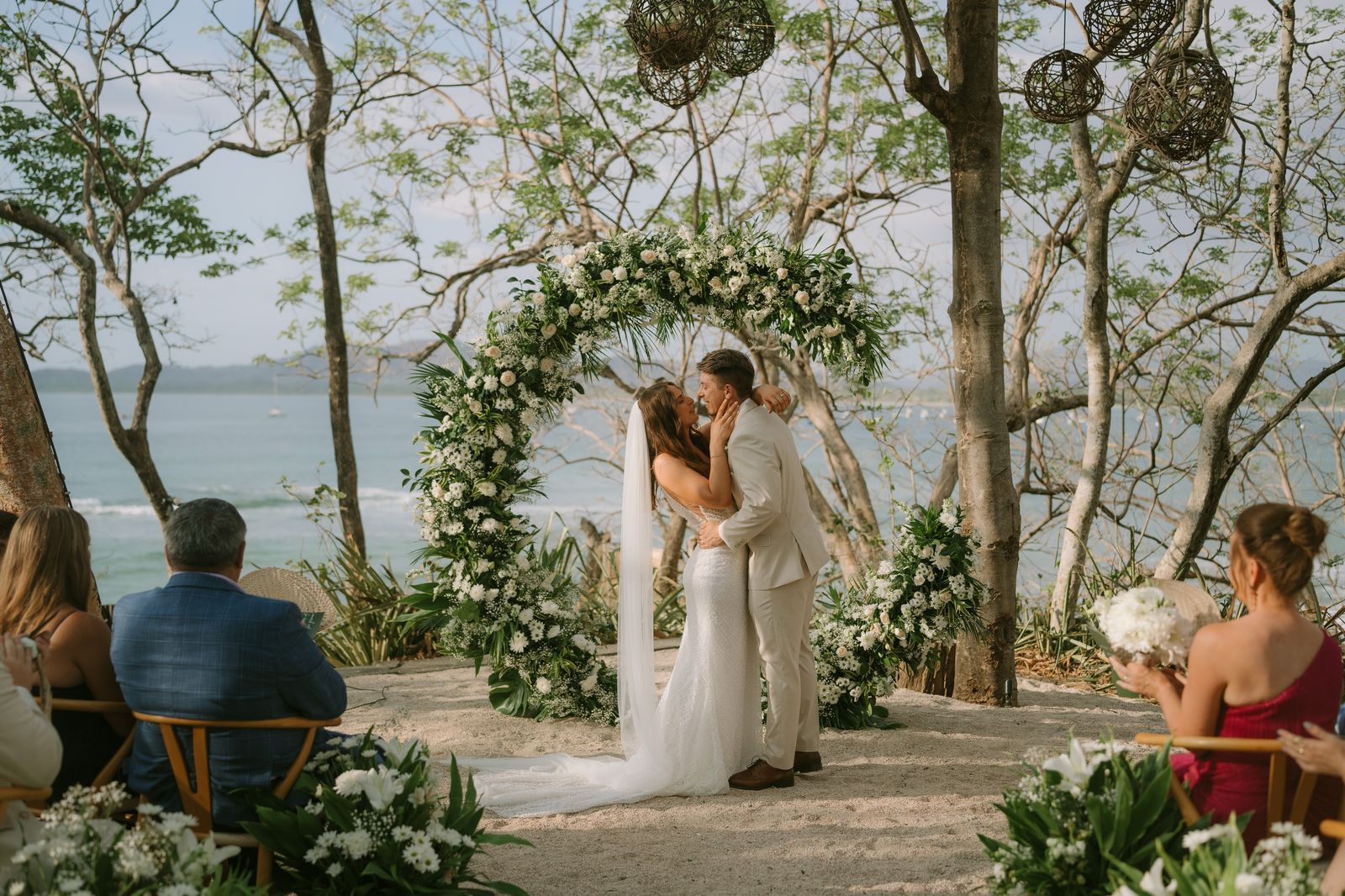 A bride and groom are kissing during their wedding ceremony in front of the ocean.