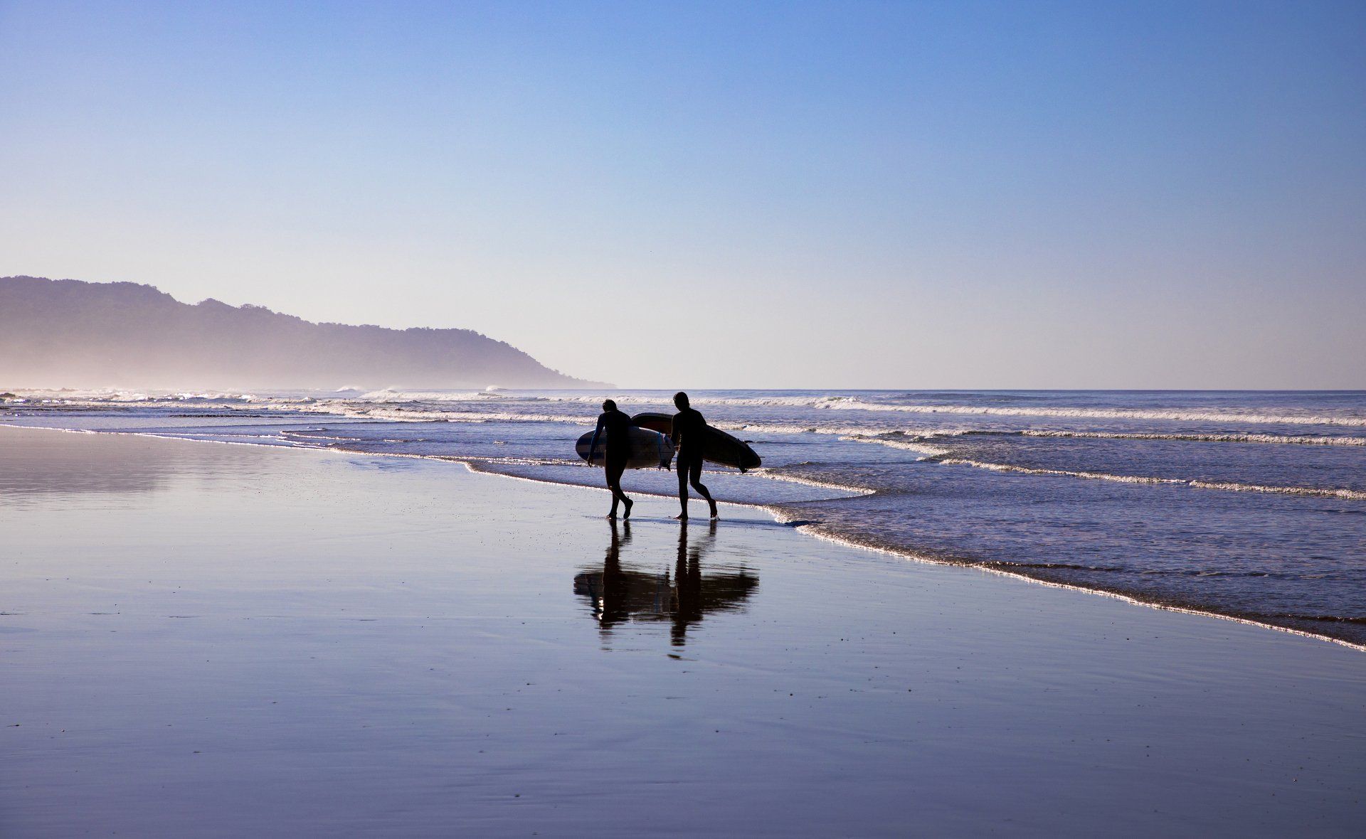Two surfers are walking along the beach carrying their surfboards.