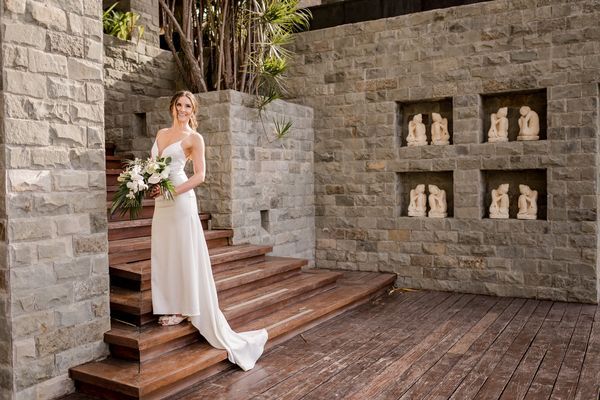 A bride in a white dress is standing on a set of wooden stairs holding a bouquet of flowers.
