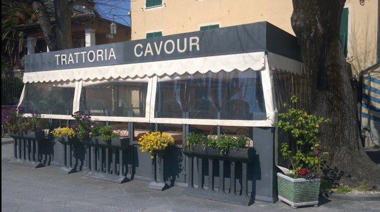 La vetrina della Trattoria Cavour è dotata di tenda a righe, vasi di fiori e tavolini all'aperto sul marciapiede.
