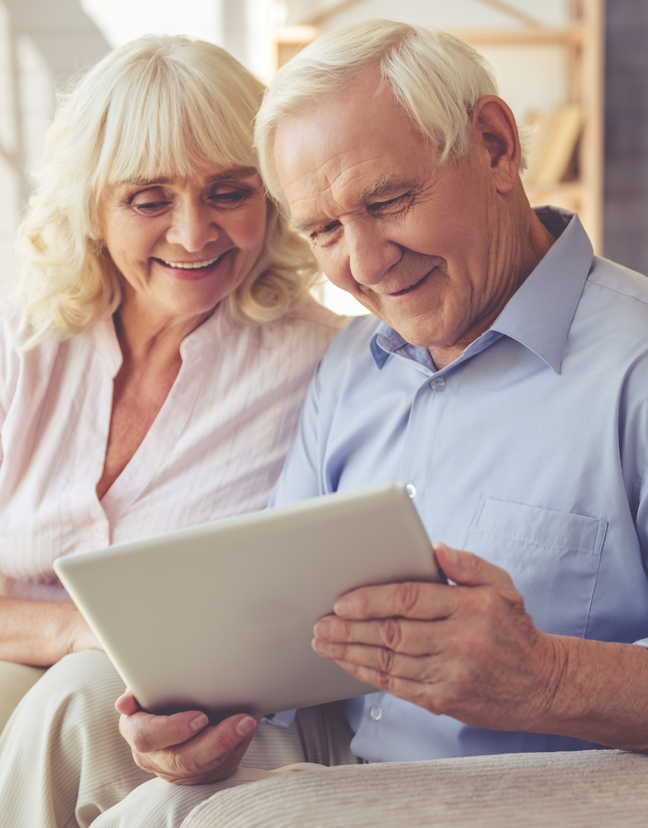 An elderly couple is sitting on a couch looking at a tablet computer.
