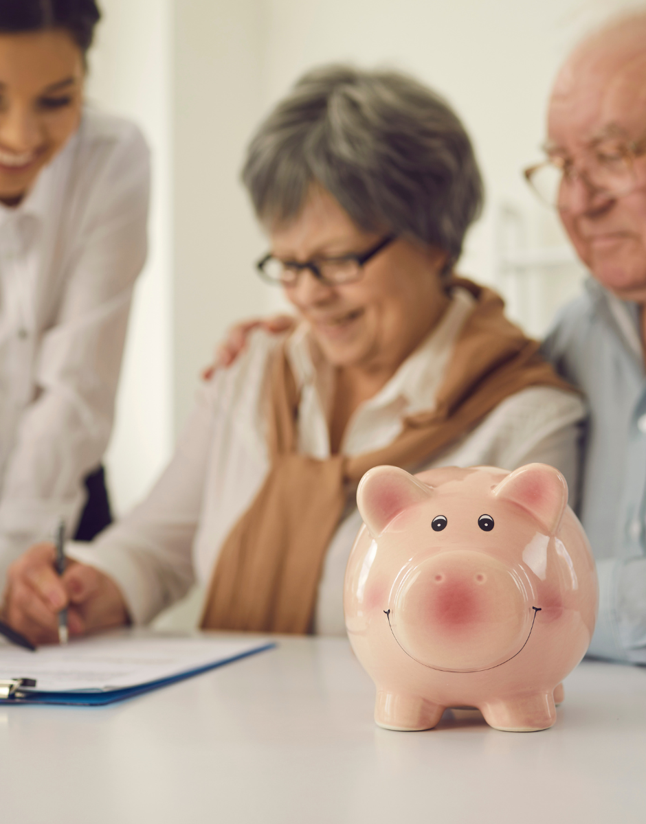 An elderly couple is signing a document with a piggy bank in the foreground.