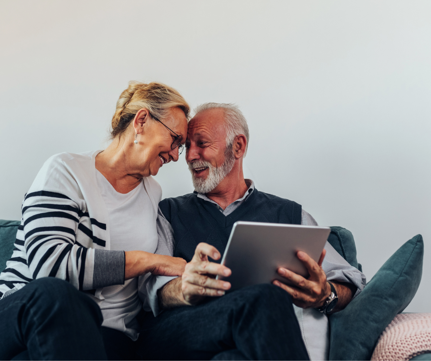 An elderly couple is sitting on a couch looking at a tablet.