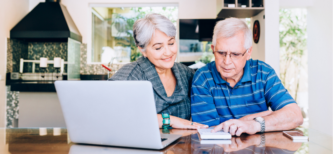An elderly couple is sitting at a table looking at a laptop computer.