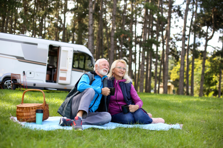 An elderly couple is sitting on a blanket in front of a rv.
