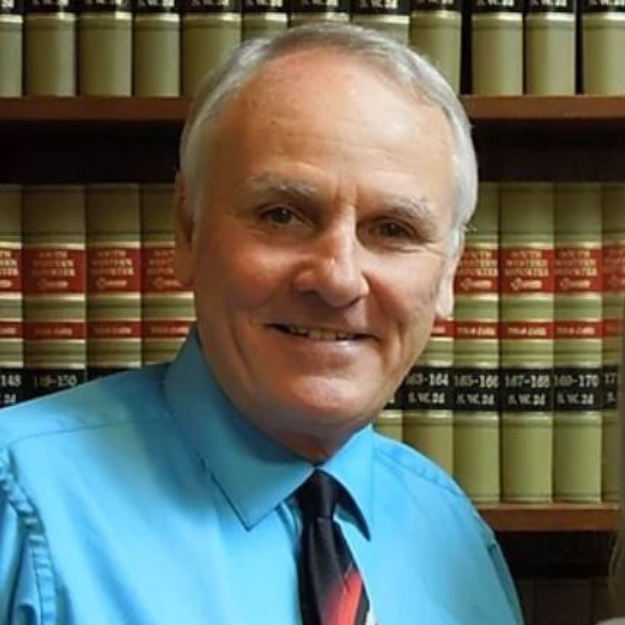 A man in a blue shirt and tie is smiling in front of a bookshelf