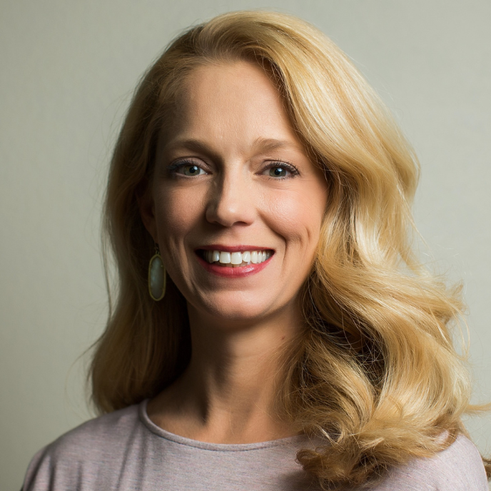 A woman with blonde hair is smiling and wearing a pink shirt.