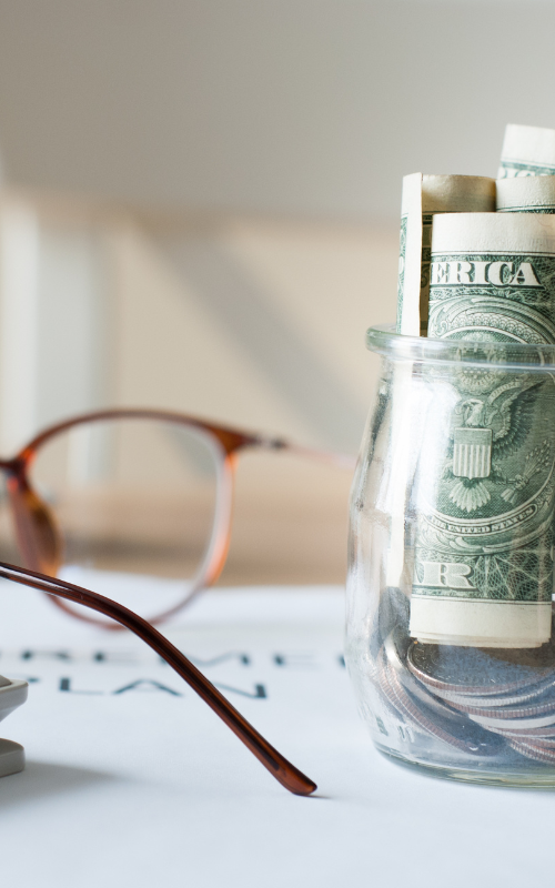 A glass jar filled with money next to a pair of glasses.