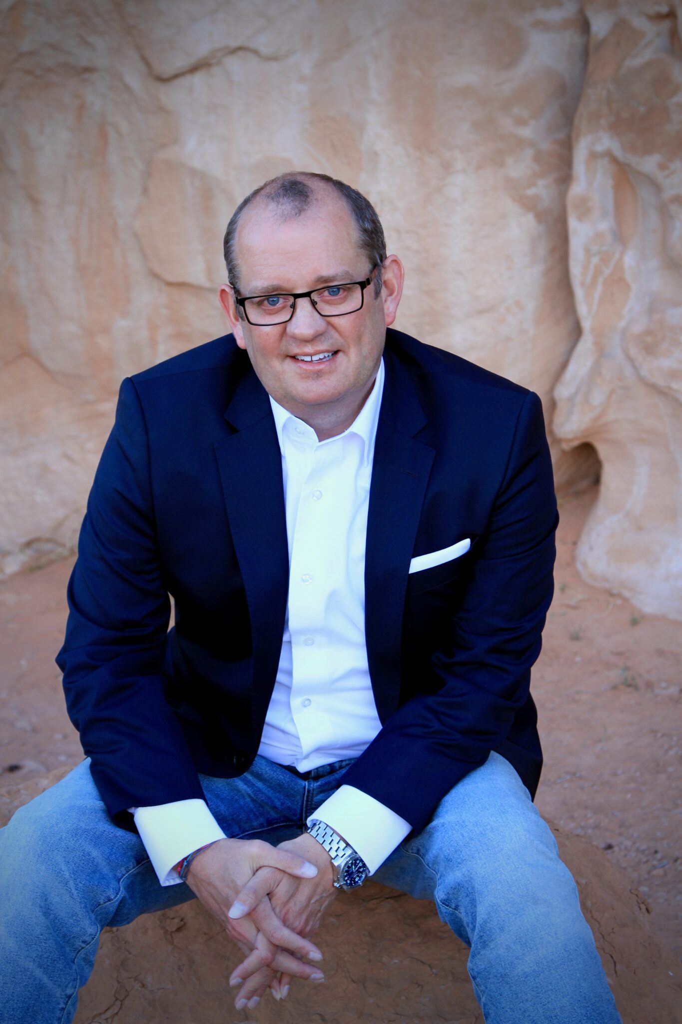 Man seated on a rock, smiling in a navy blazer and jeans against a tan stone wall.