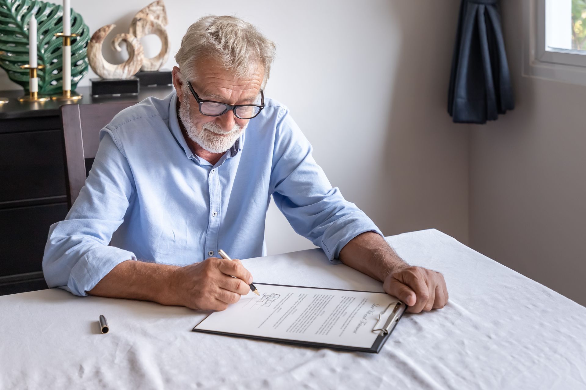 Man writing on a document at a table in a bright room, wearing glasses and a light blue shirt