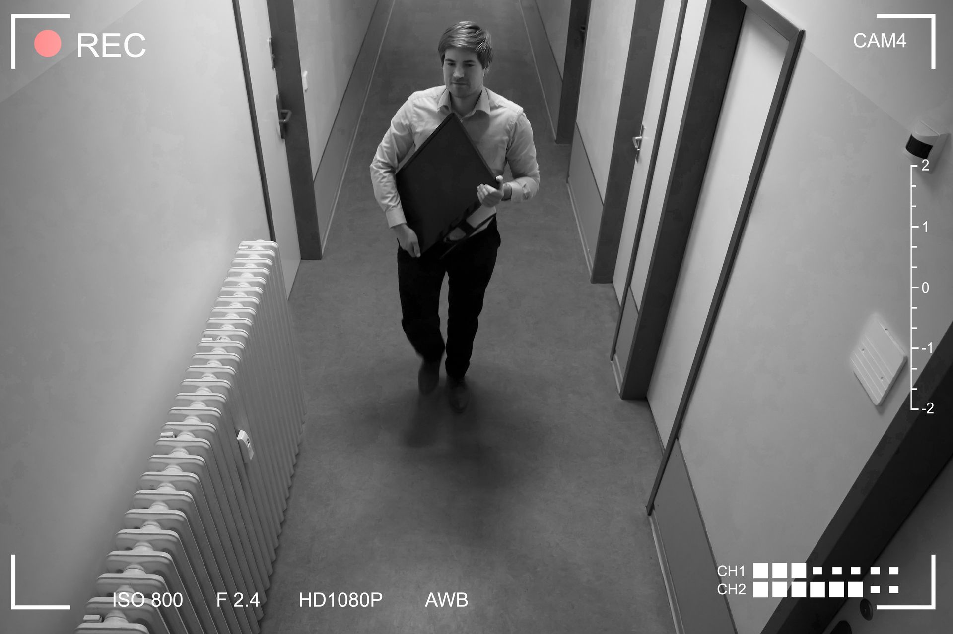 Person walking down a narrow hallway carrying a laptop under security camera view.
