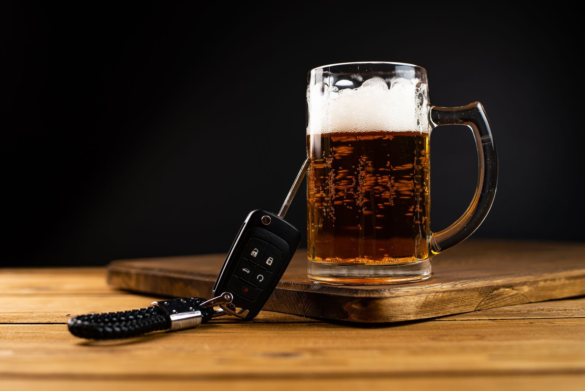 A beer mug on a wooden table beside car keys, with a dark background.