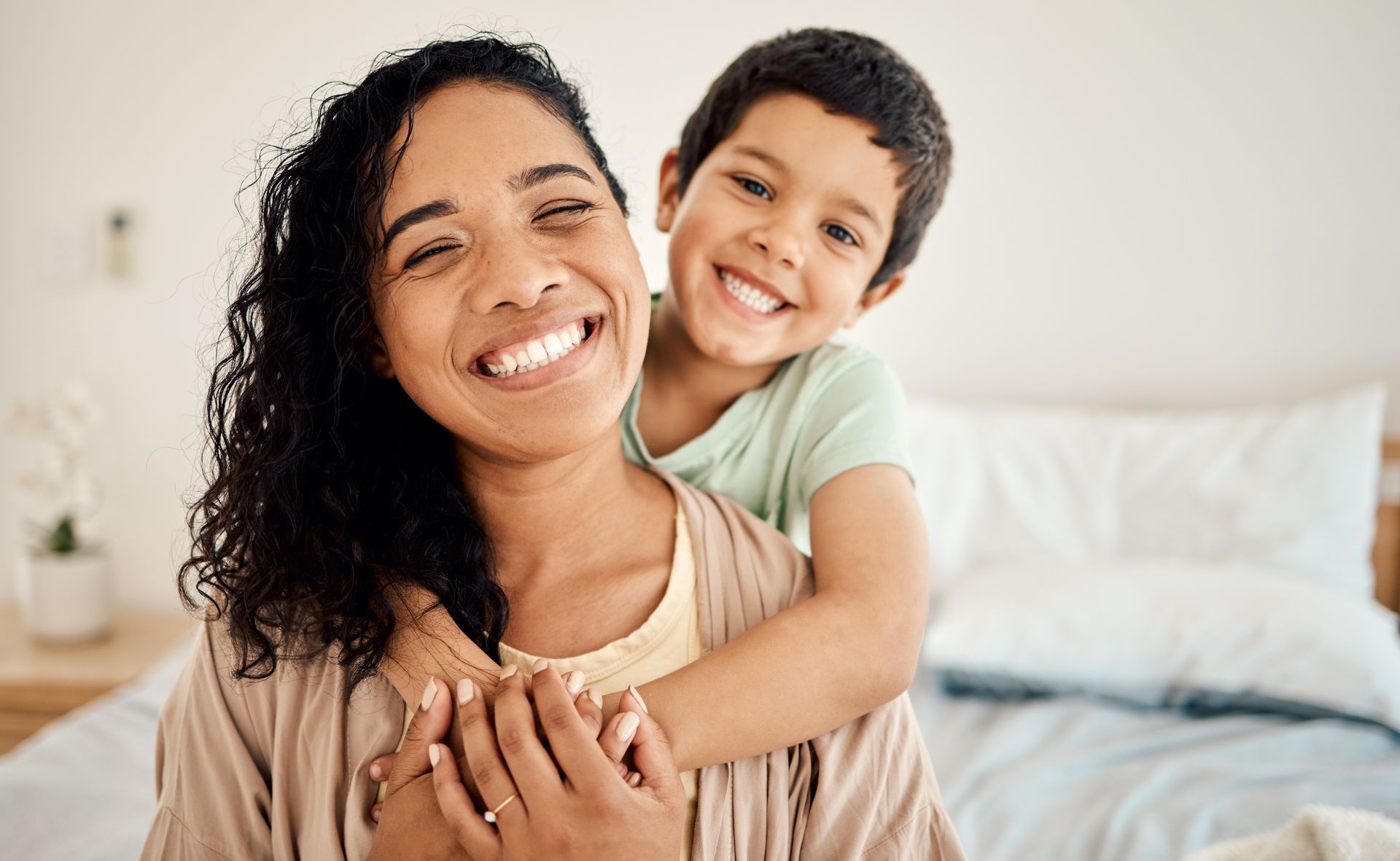 Smiling parent with child in a bright bedroom, child hugging from behind