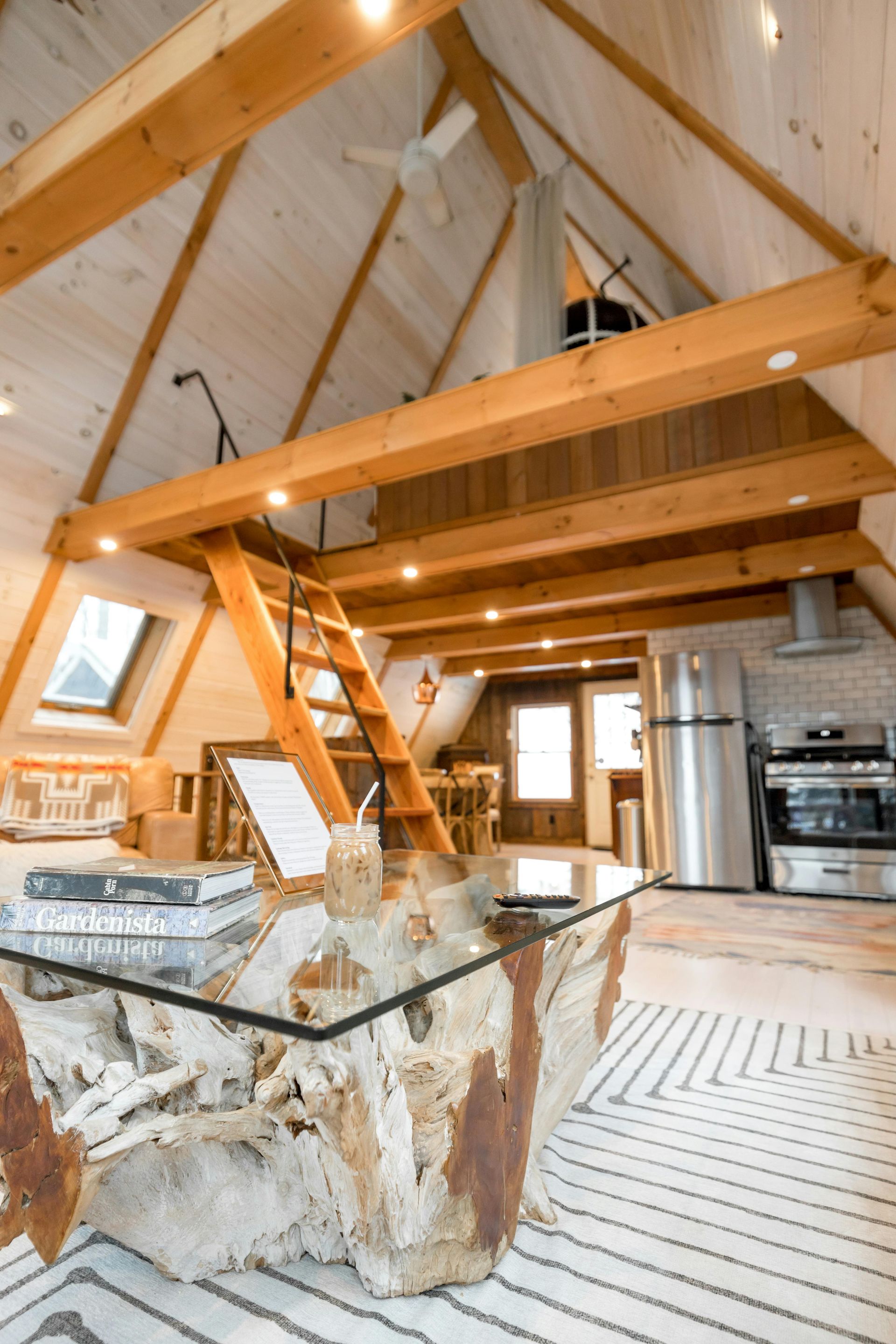 Rustic loft kitchen with wooden beams, ladder, glass table, and stone island beneath a high vaulted ceiling