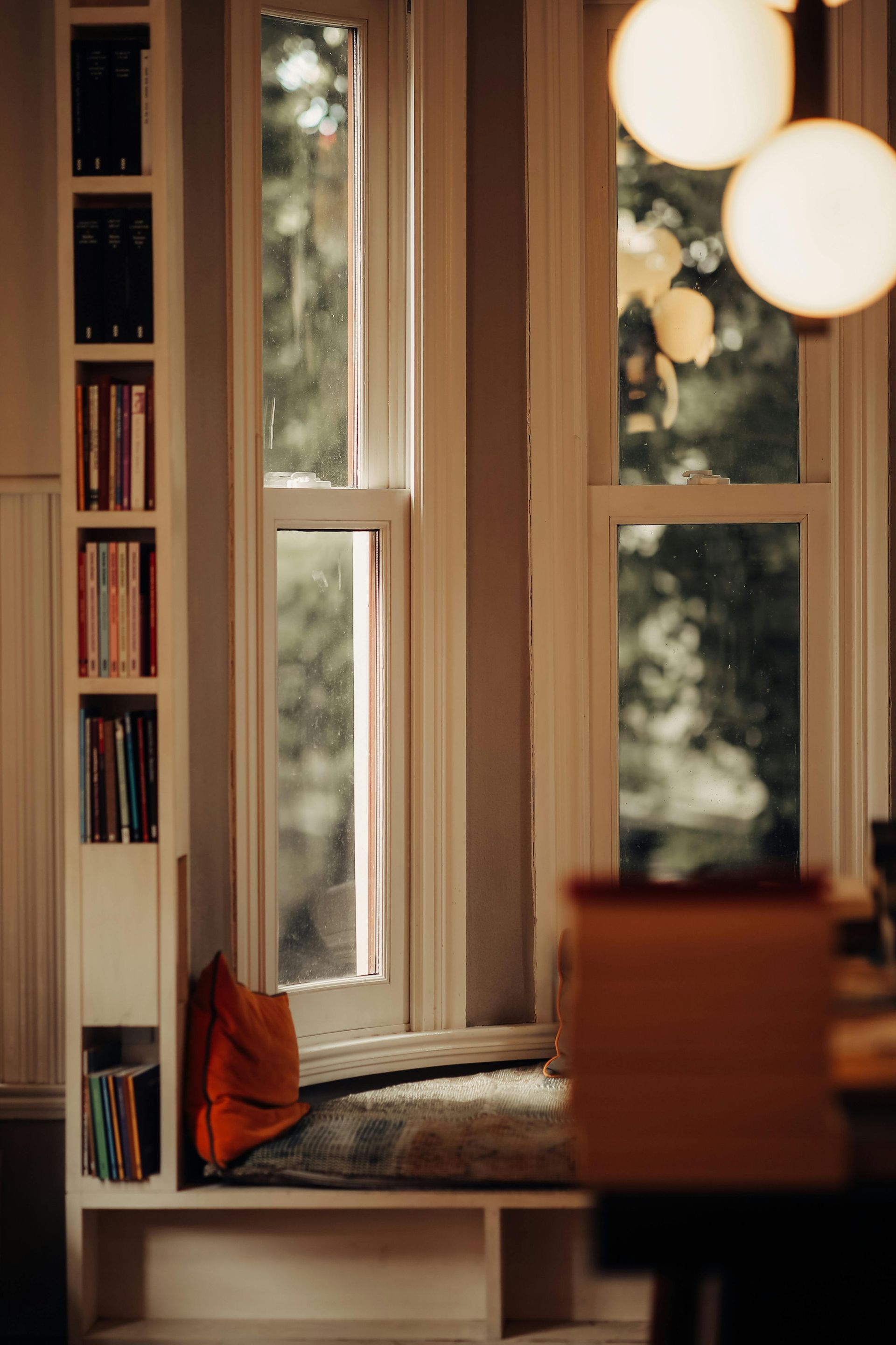 Cozy reading nook with a window seat, bookshelves, and warm pendant lights overlooking trees outside