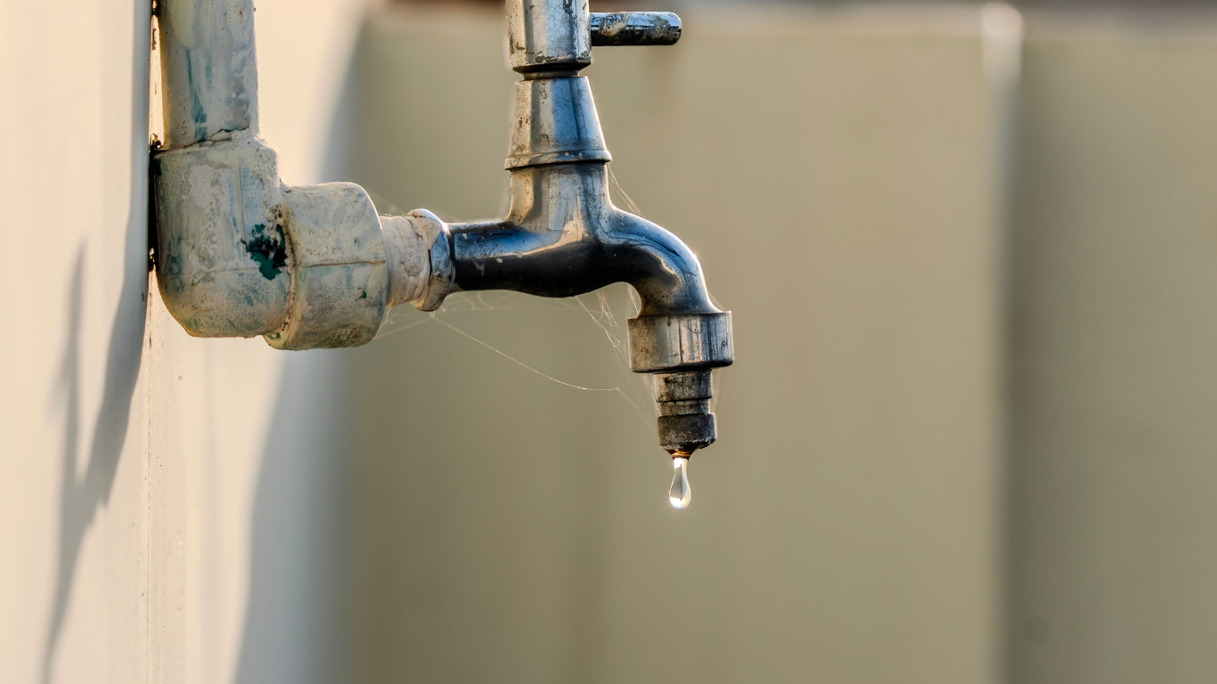 Leaky outdoor metal faucet with a water droplet against a blurred beige wall