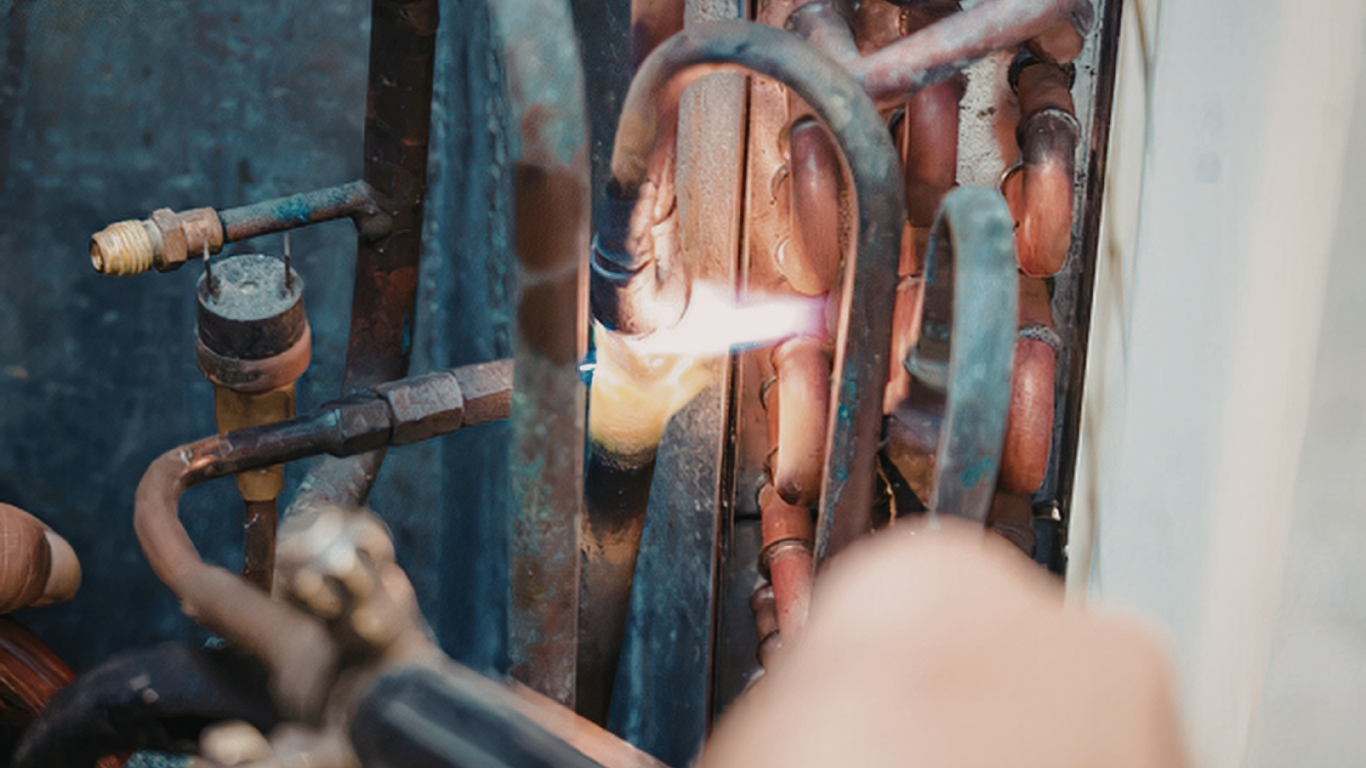 Technician welding copper pipes inside an HVAC unit, with bright sparks and blue-black tubing.