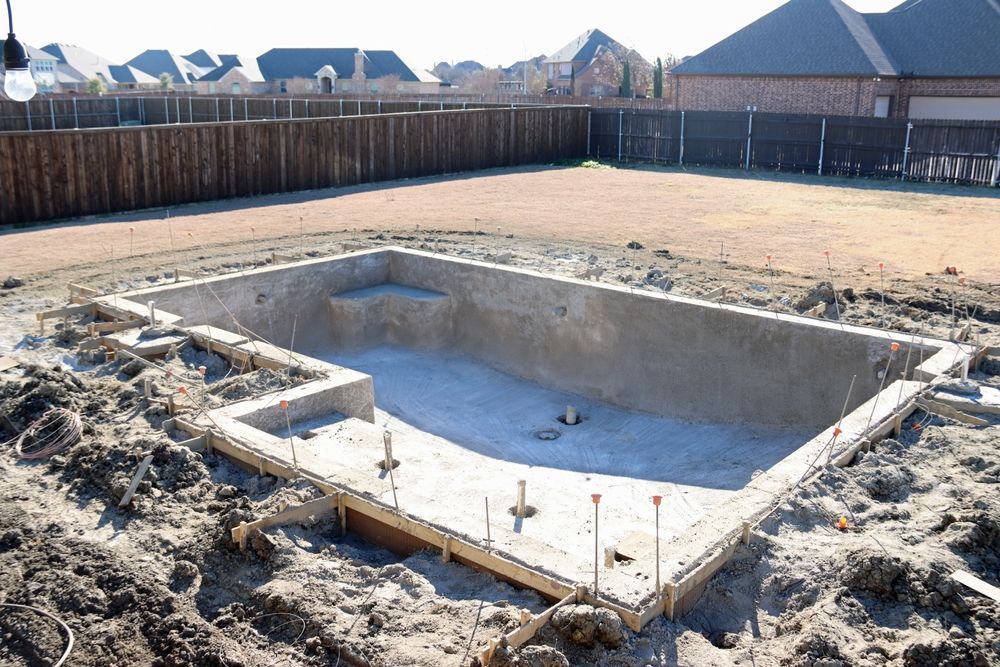 Shallow concrete foundation pit under construction in a fenced residential lot, with houses in the background