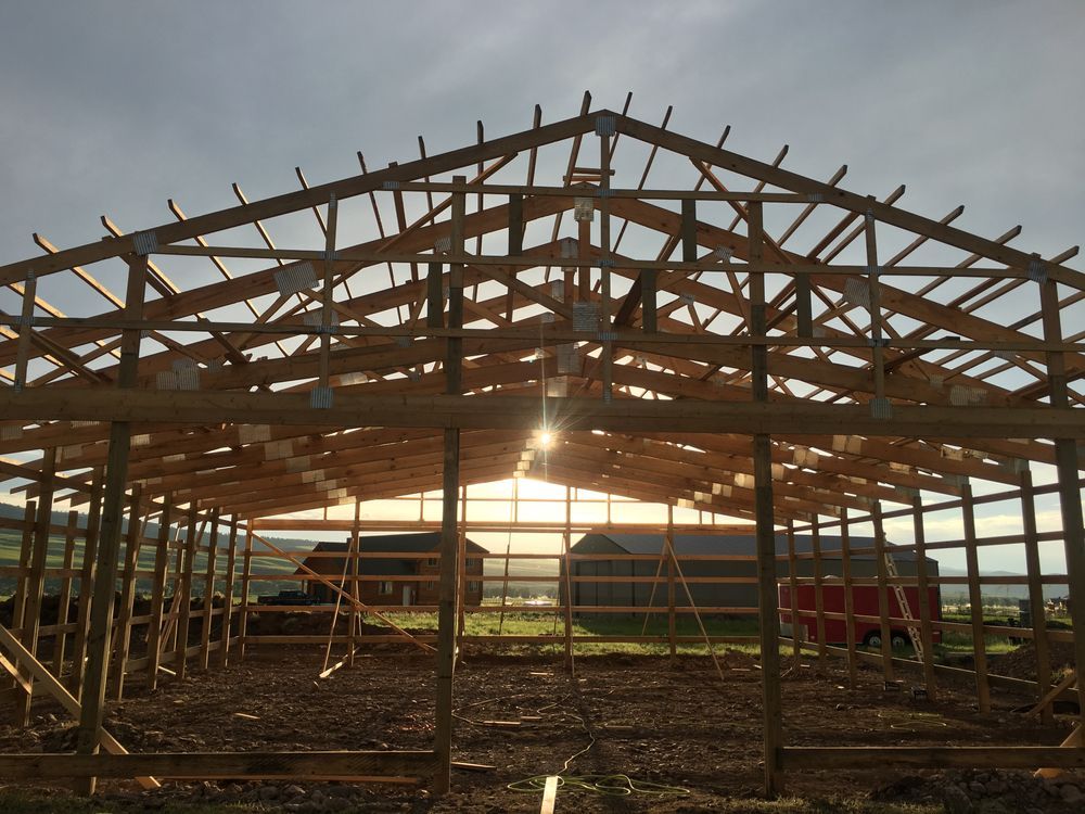 Wooden barn frame under construction at sunset, with beams silhouetted against the sky.