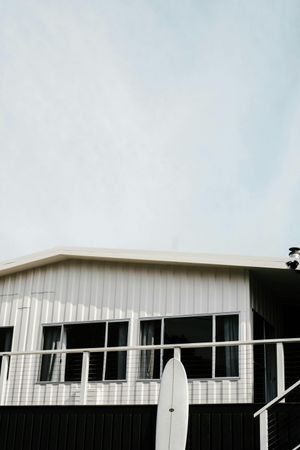 White beach house with a surfboard leaning on the railing, clear sky.