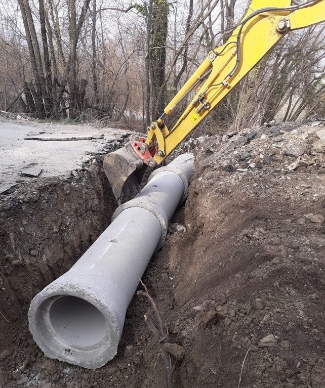 Un escavatore giallo sta posizionando un grande tubo di cemento in una trincea. La trincea è circondata da terra, con alberi sullo sfondo.