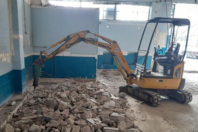 Un escavatore giallo con un martello pneumatico demolisce il cemento all'interno di un edificio con pareti blu.