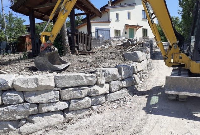 Escavatore giallo che costruisce un muro di contenimento in pietra vicino a una casa.
