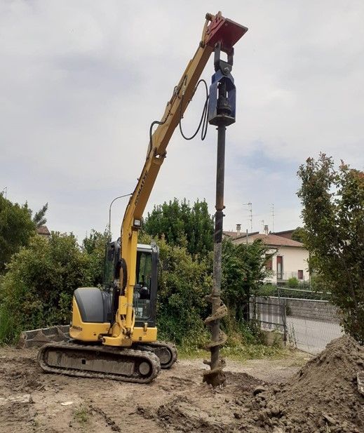 Un escavatore giallo sta perforando un buco nel terreno; sullo sfondo si vede una zona residenziale.