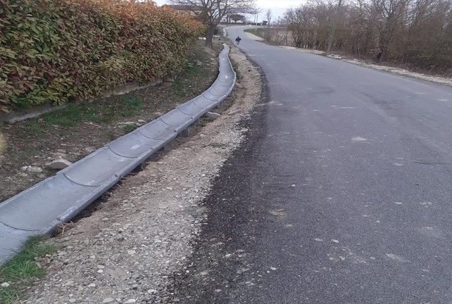 Strada asfaltata con un canale di drenaggio in cemento lungo un terrapieno fiancheggiato da cespugli.